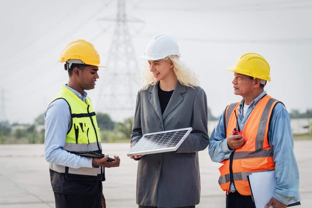 Three people, two men wearing hard helmets and vest and one woman wearing hard helmet, are standing outside, the woman is holding a solar panel